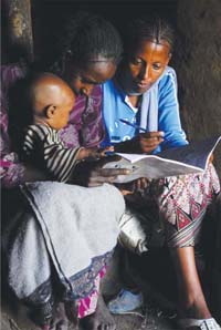 Two women a re looking through a workbook. A small child sits on one of the women's lap.