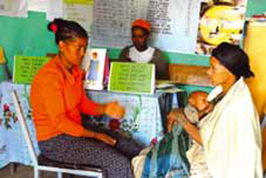 A health professional is talking to a mother. A baby lays asleep on the mothers lap.