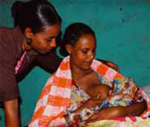 A mother is breastfeeding her newborn baby whilst a health professional watches over her shoulder.