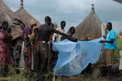 A group of people are gathered outside their homes demonstrating how to use the insecticide-treated bed nets. A woman and child are seated underneath it.