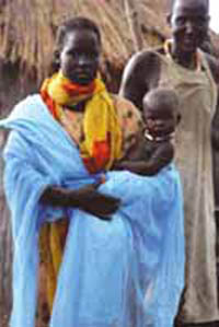 A woman standing outside her home. She is holding her baby and has an insecticide-treated net wrapped around her.